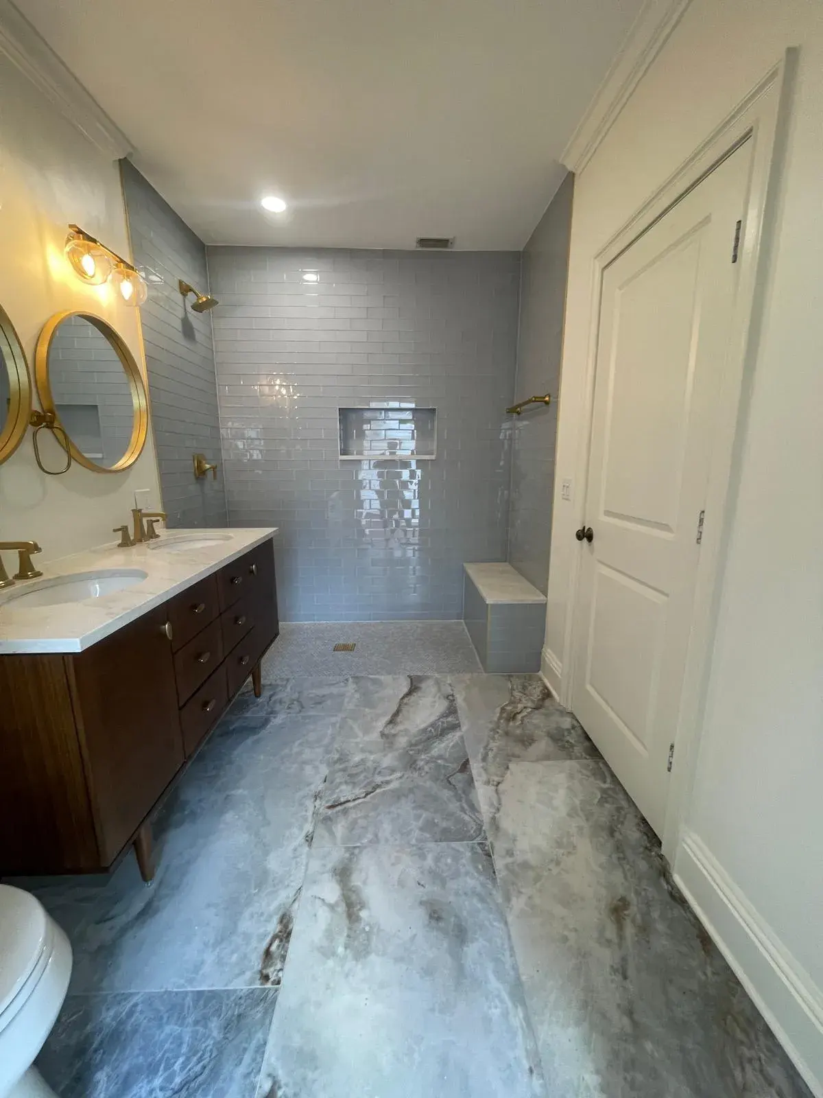 Long bathroom with a wood double vanity, two round mirrors, gold fixtures and marble-look floor tile