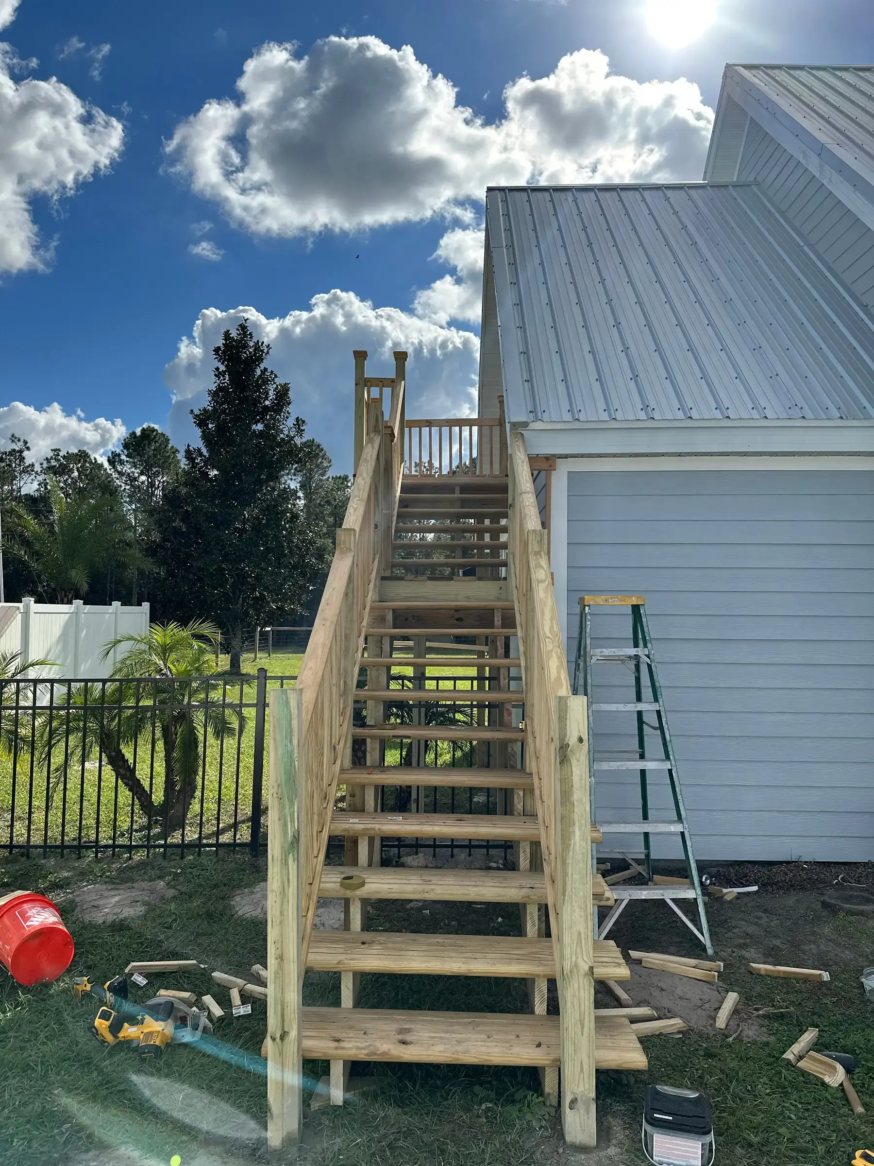 Wood exterior staircase under construction beside a house, with lumber offcuts and a ladder on the grass