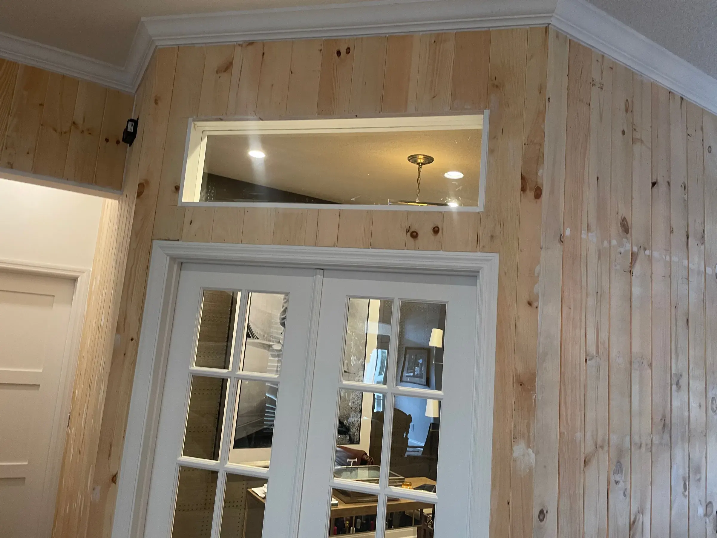 Natural wood wall paneling around white French doors with a transom window and crown molding