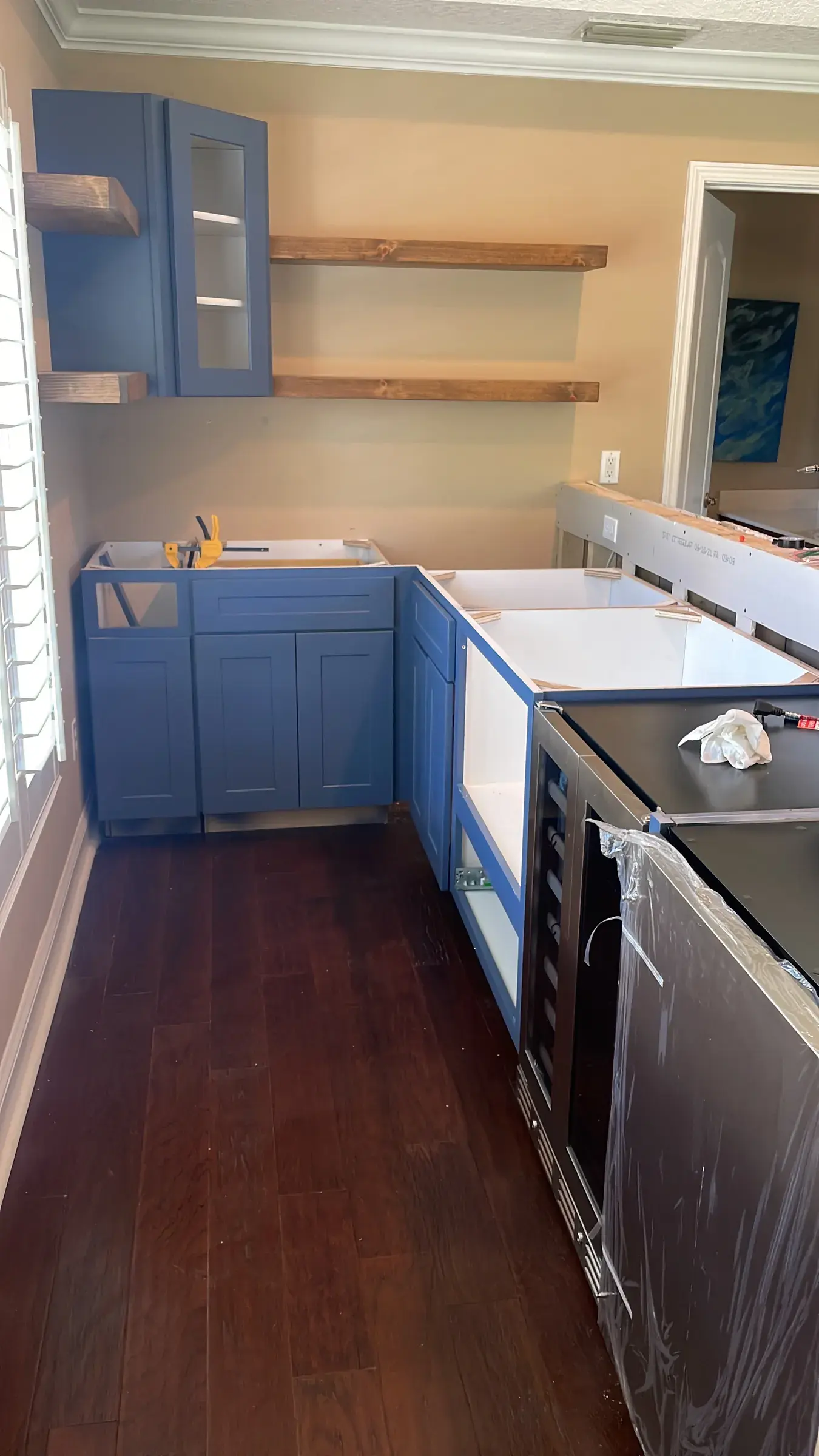 Blue lower cabinets and open wood shelves during installation, beside appliances wrapped in plastic and dark wood flooring