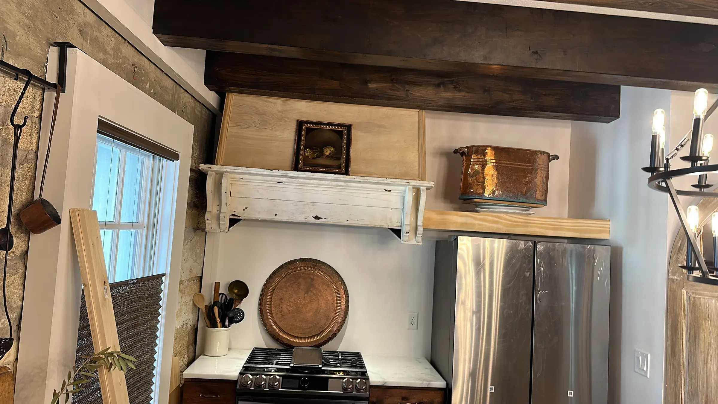 Kitchen range below a white mantel-style hood, with dark wood ceiling beams, a copper pot on a wood shelf and a stainless refrigerator