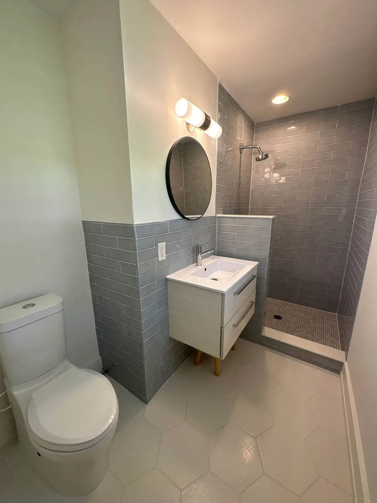 Bathroom with gray tile, a vanity with legs, a round mirror, vanity lighting and a tiled shower.